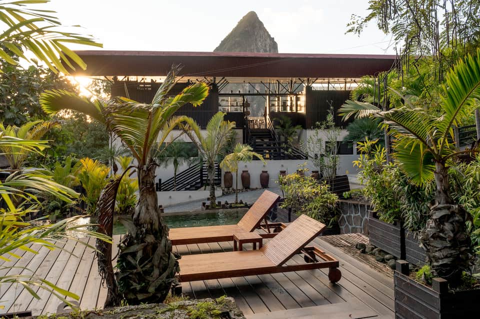 Tropical resort courtyard with loungers on wooden deck, lush palm plants, and mountain backdrop