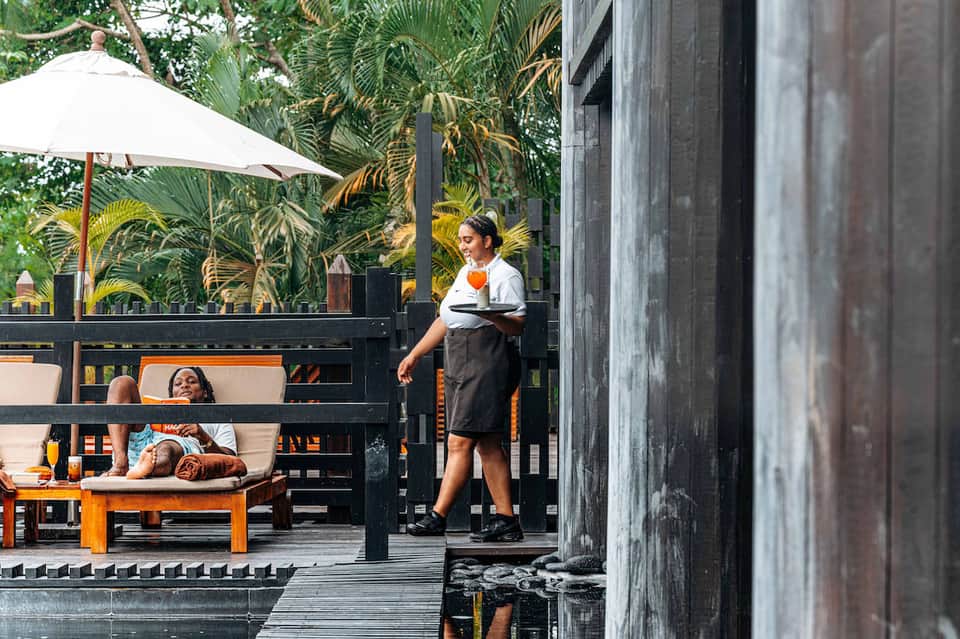 Beachfront restaurant patio with server, white umbrella, lounging guests, and tropical plants