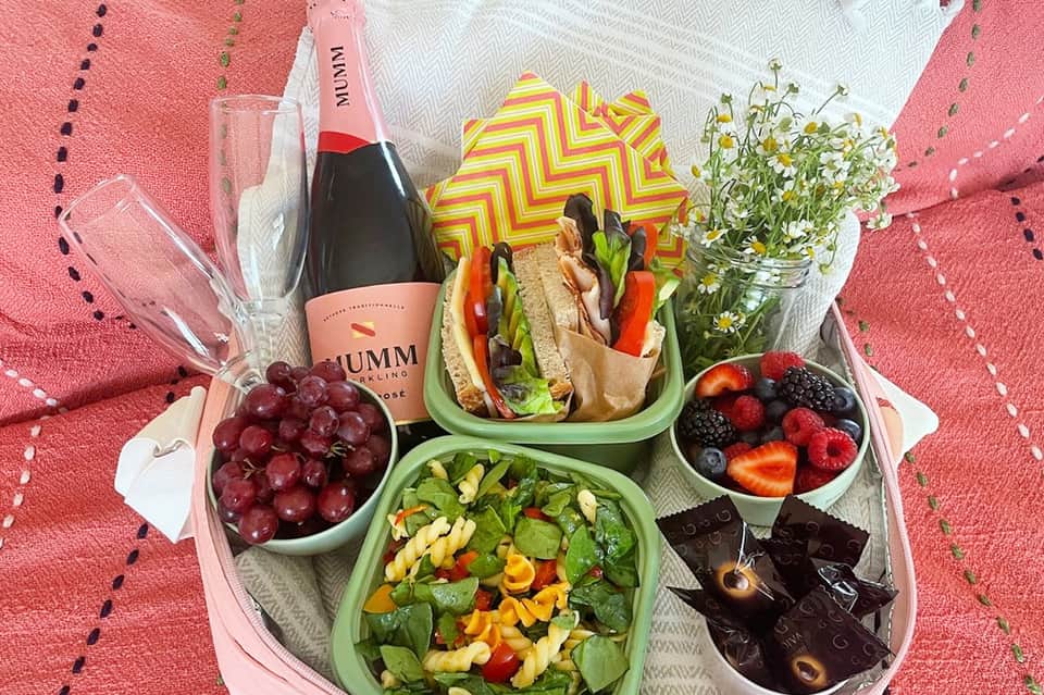 Gourmet picnic spread with champagne, sandwiches, fresh berries, salad, and flowers on pink blanket