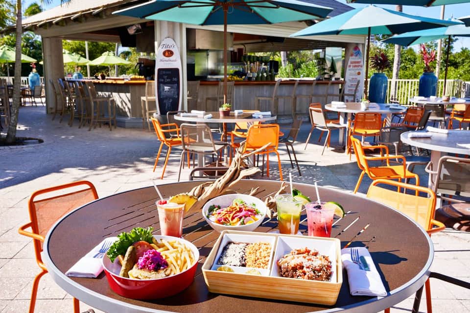 Outdoor beachfront restaurant with orange chairs, colorful dishes, tropical flowers, and ocean view