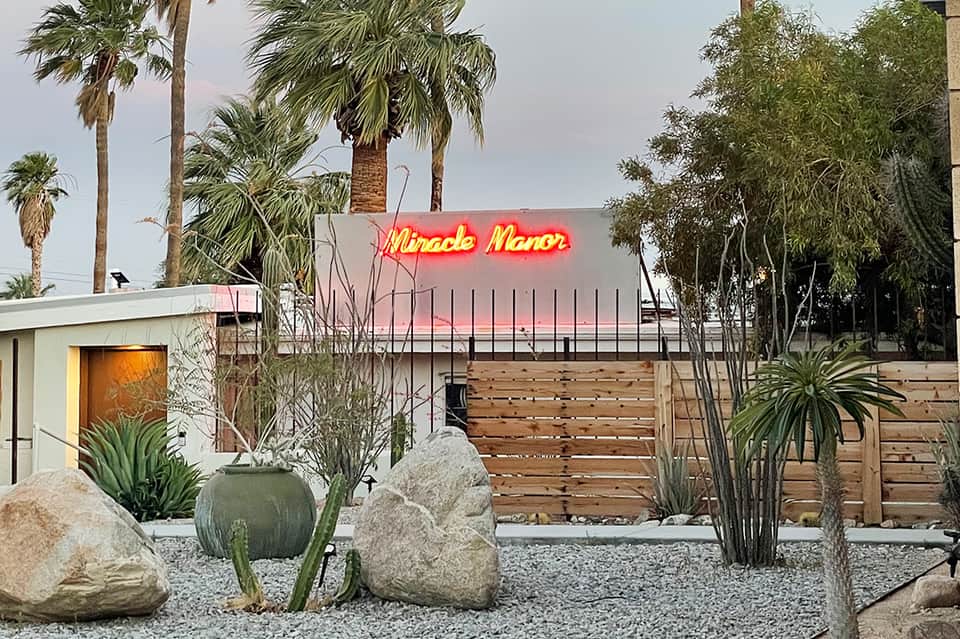 Neon 'Miracle Manor' sign on mid-century modern building with palm trees