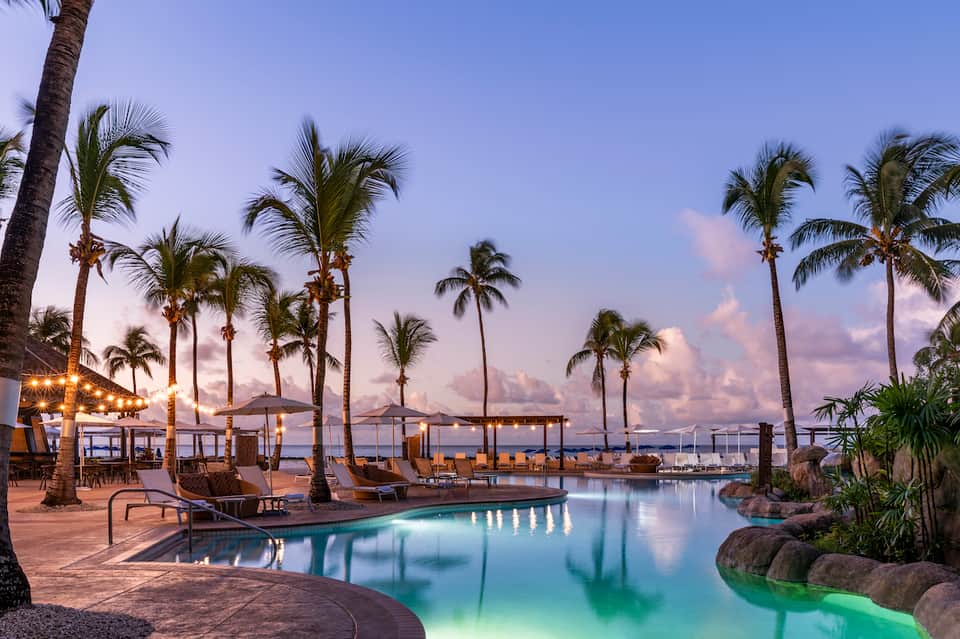 Waterfront resort pool at twilight surrounded by palm trees with ocean views and lounge chairs
