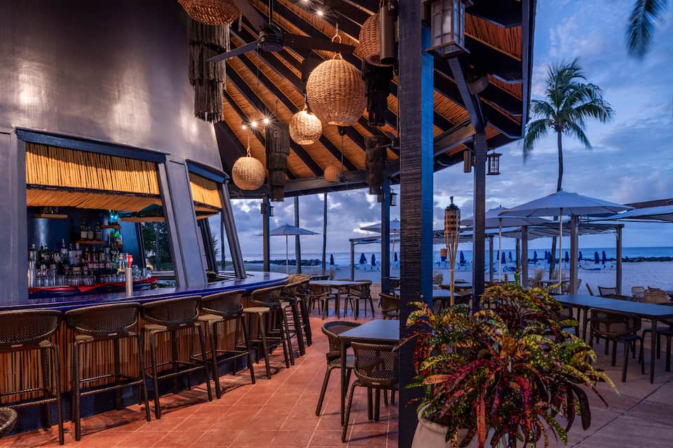 Beachfront bar at twilight with woven pendant lights, ocean view, and palm trees