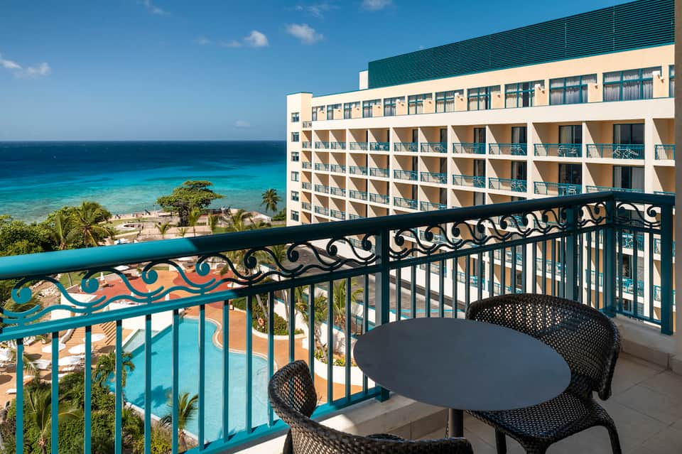 Resort balcony with ornate railing overlooking tropical pool, beach, and ocean with palm trees