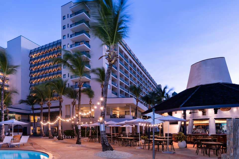 Resort beachfront at dusk with palm trees, lounge seating, and illuminated building