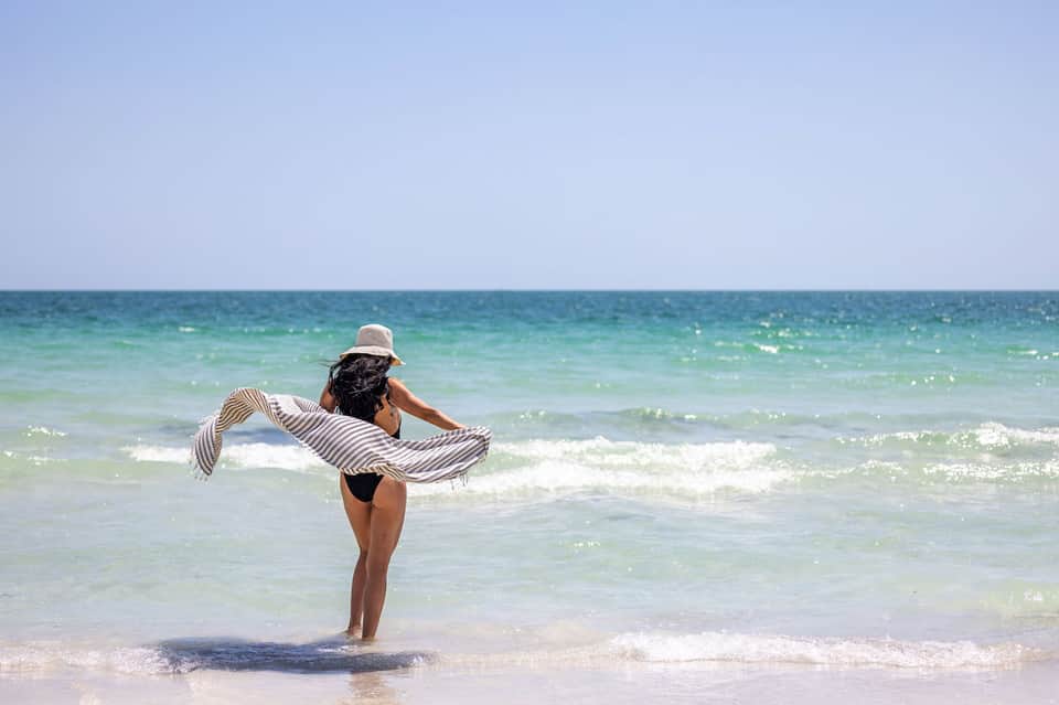 Woman in straw hat wading in turquoise ocean waves on sandy beach