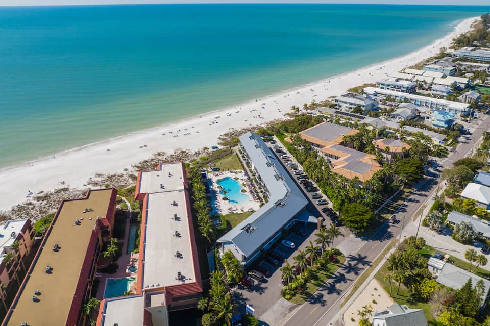 Aerial view of beachfront resort with pool, sandy beach, palm trees, and surrounding accommodations
