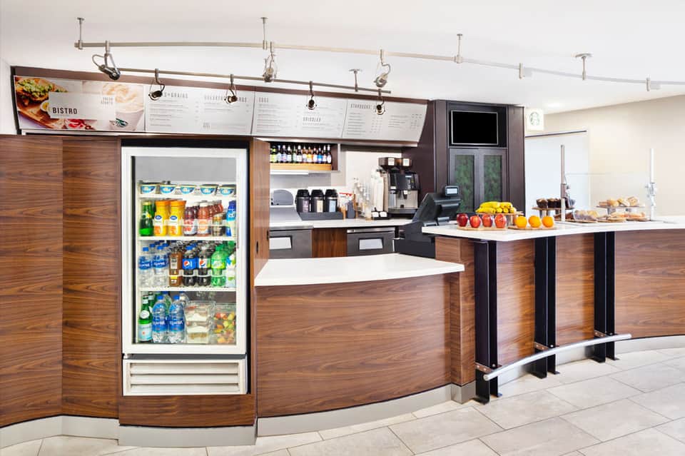 Modern hotel café counter with wood paneling, refrigerated beverage display, menu boards, and service area
