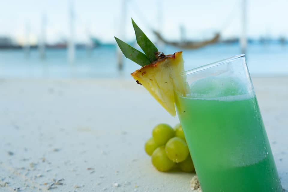 Tropical beach cocktail with pineapple garnish and green liquid on white sand with sailboats