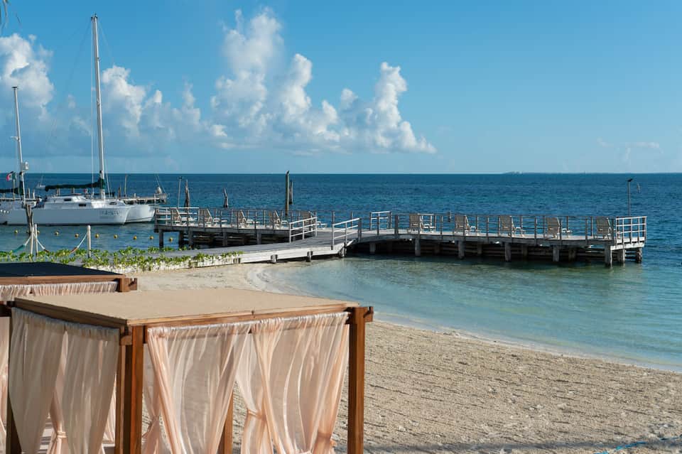 Tropical beach scene with sailboat at dock, white sand, palm trees, and calm turquoise water.