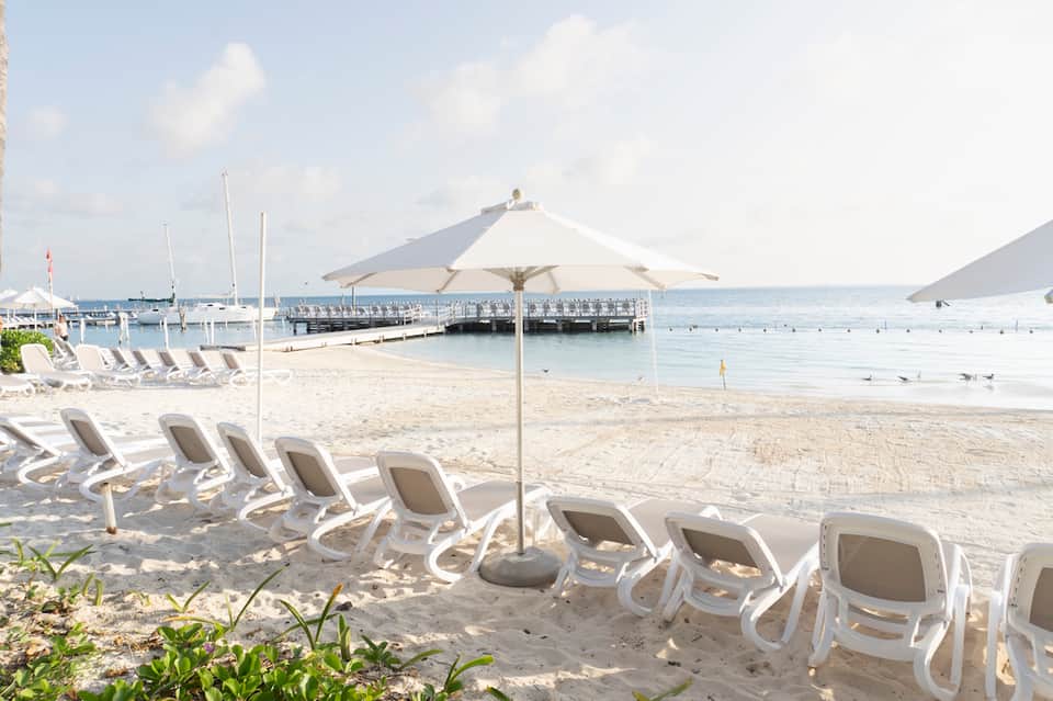 White lounge chairs with umbrellas on sandy beach with boats and pier in background