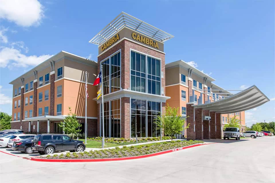 Modern hotel exterior with brick and orange facade, central tower marked Cambria, and parking lot