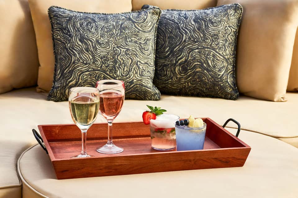 Luxurious poolside setup with decorative pillows, cocktails on wooden tray, and cream cushioned seating
