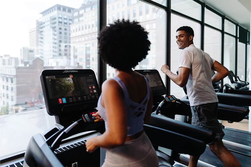 Two people exercising on treadmills in a gym with floor-to-ceiling windows overlooking city skyline