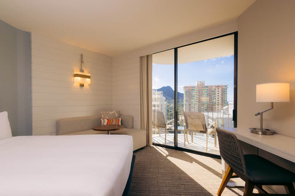 Hotel bedroom with queen bed, work desk, and balcony overlooking mountains and city buildings in daylight