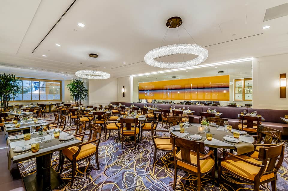 Upscale dining room with round tables, gold pendant chandeliers, and sunset artwork on wall