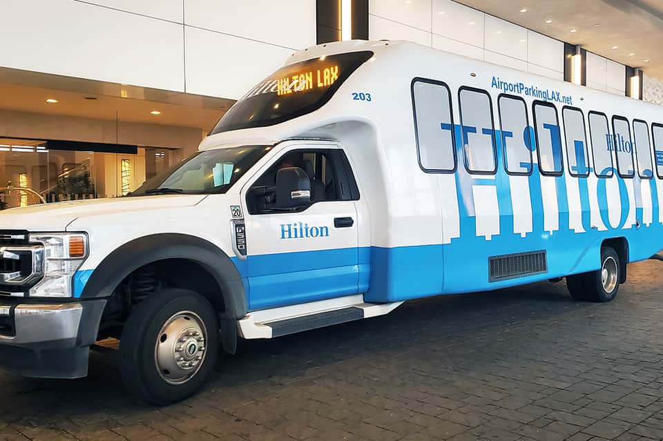 White and blue Hilton airport shuttle bus with large passenger windows parked in hotel entrance