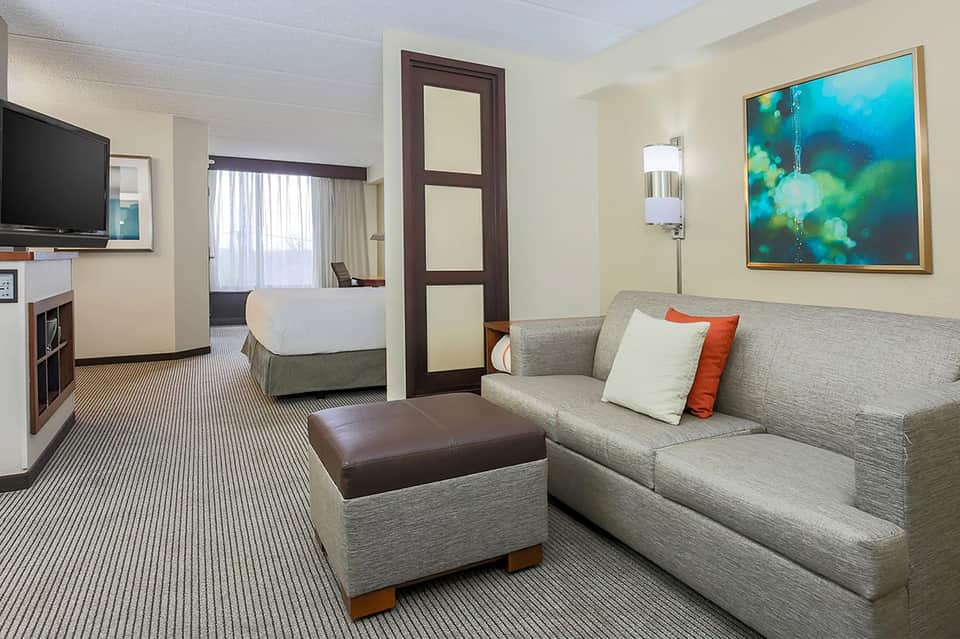 Hotel suite with queen bed, sofa, TV, and warm neutral décor with accent pillows