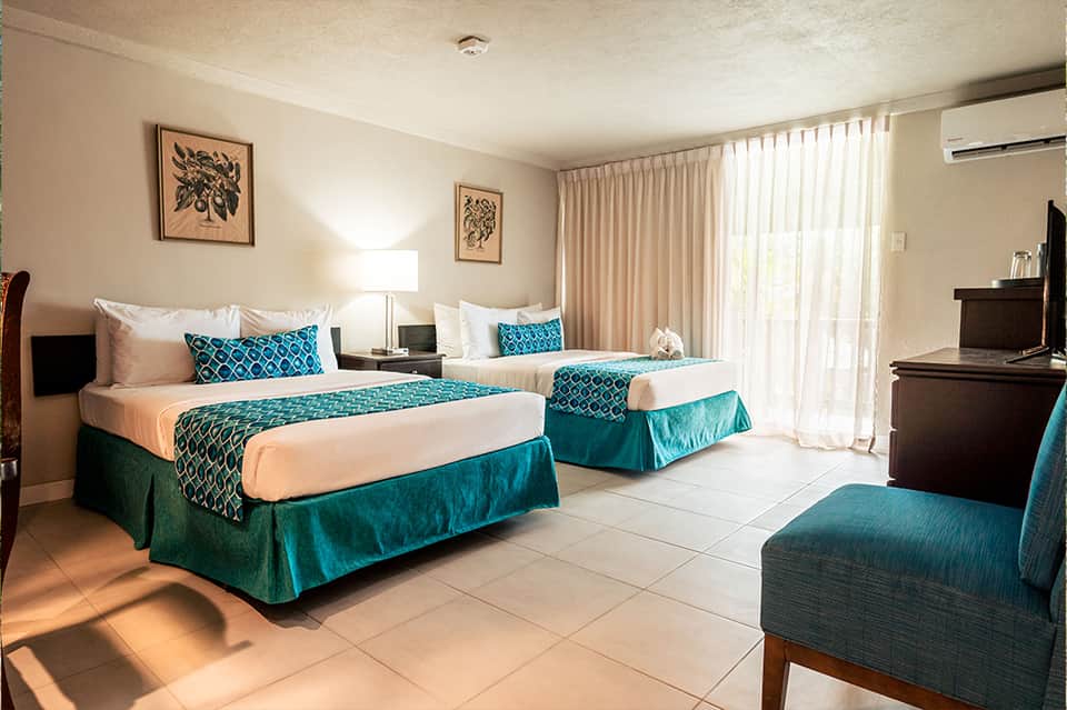 Hotel room with two queen beds, teal bedding, sitting area, and patio access with beige curtains