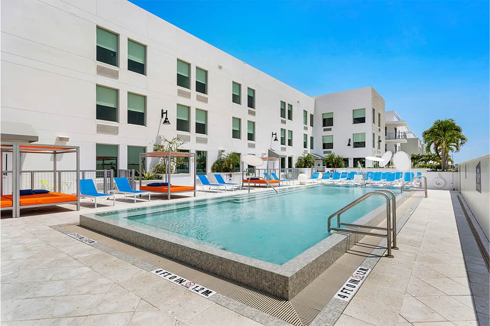 Resort pool area with blue loungers, white building, palm trees, and clear sky