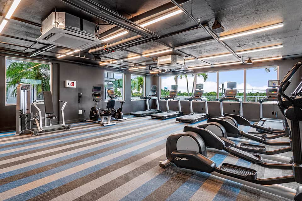 Spacious fitness center with rows of exercise equipment, large windows with ocean views, and striped blue carpet