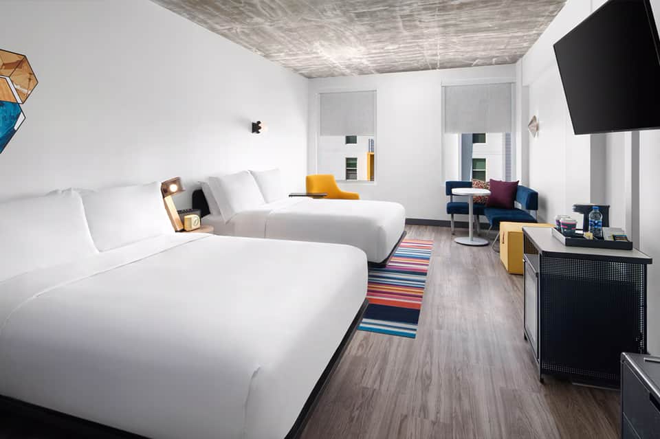 Modern hotel room with two queen beds, white bedding, flat-screen TV, and minimalist décor