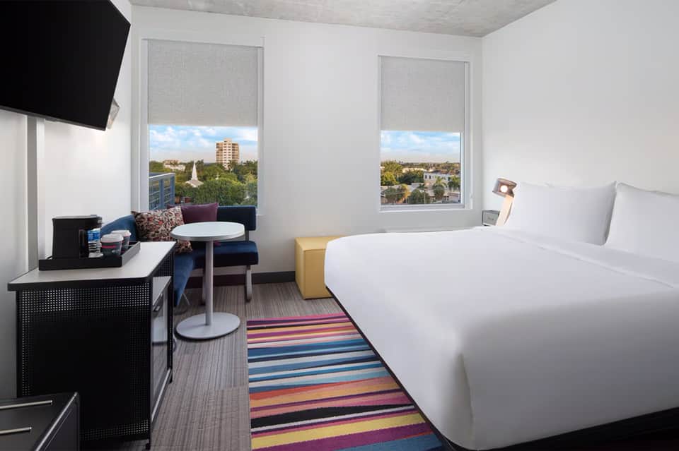 Modern hotel room with white beds, desk, city views through large windows