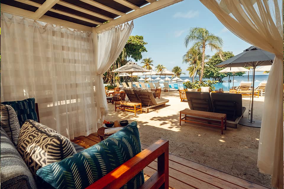 Beachfront cabana with sheer curtains, wooden furniture, umbrellas, and ocean view with palm trees