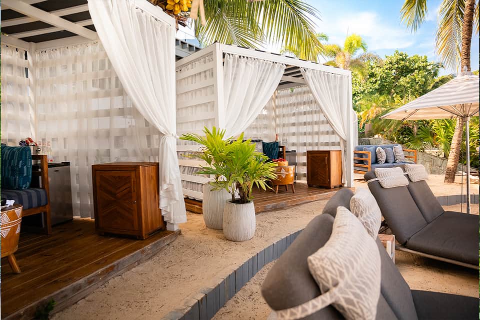Beachfront cabanas with white curtains, wooden decking, tropical plants, and lounge chairs on sand