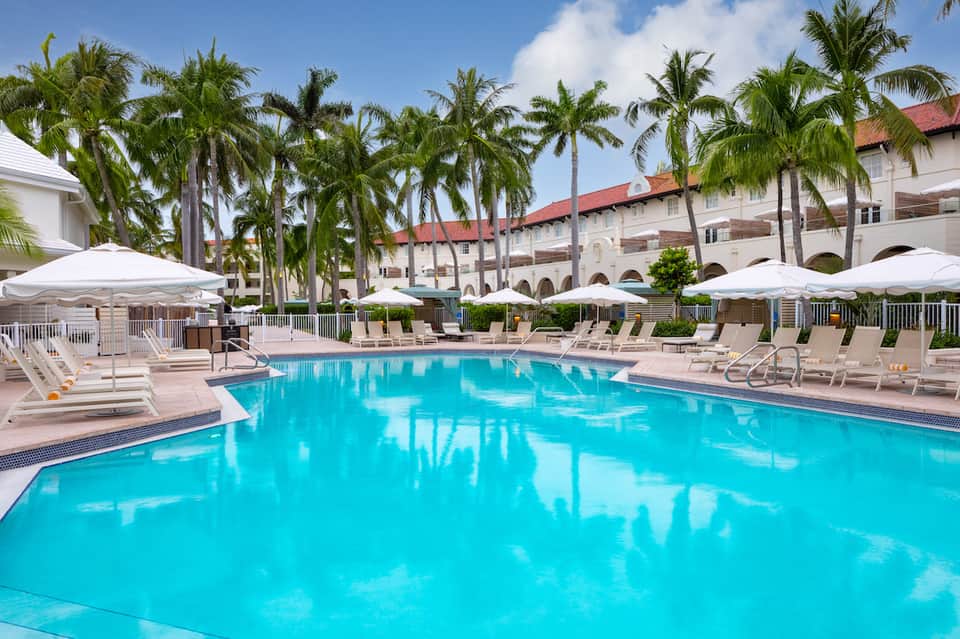 Resort swimming pool with turquoise water, lounge chairs, white umbrellas, and palm trees