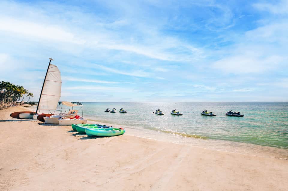 Tropical beach with sailboat, kayak, jet skis, and palm trees lining the shore