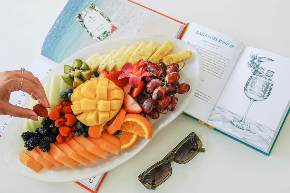 Colorful fruit and cheese platter with book and sunglasses on white table by pool