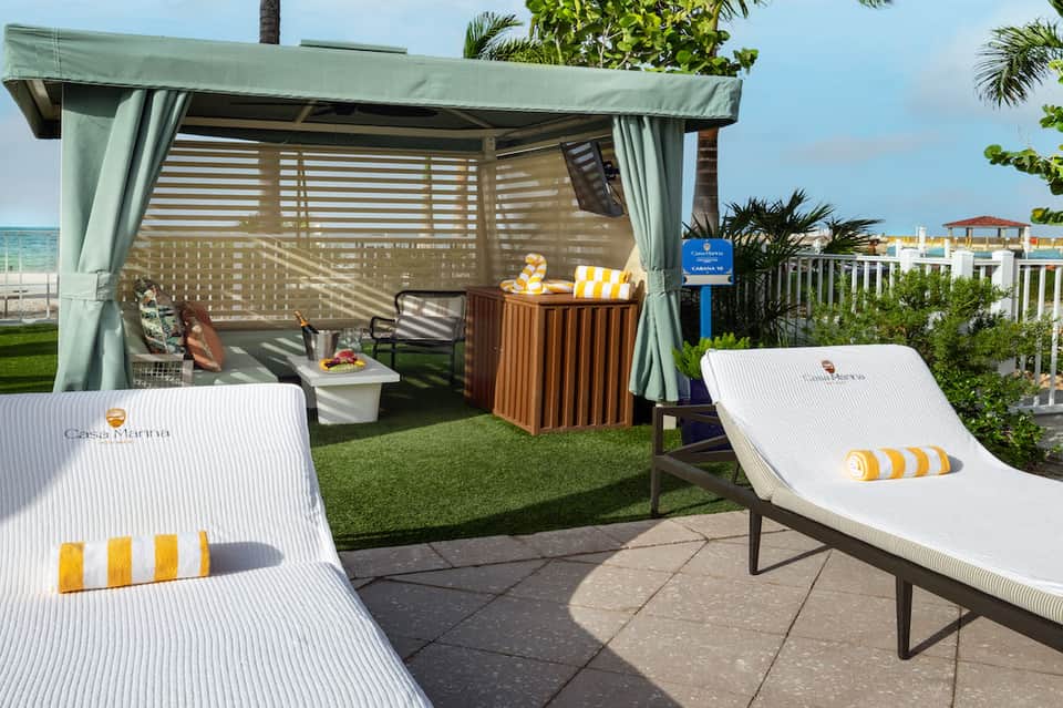 Beachfront cabana with white striped loungers, turquoise curtains, wooden bar, and ocean view
