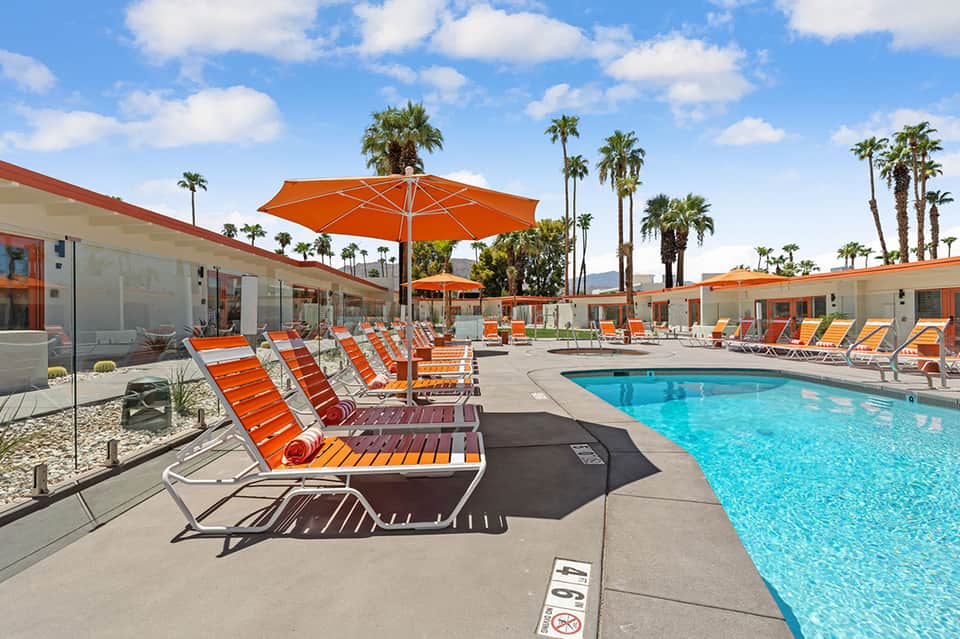 Outdoor pool area with orange and white striped lounge chairs, orange umbrellas, palm trees, and desert landscape
