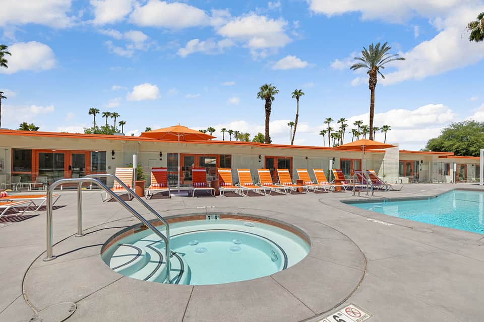 Resort pool area with hot tub, orange lounge chairs, palm trees, and mid-century modern building architecture