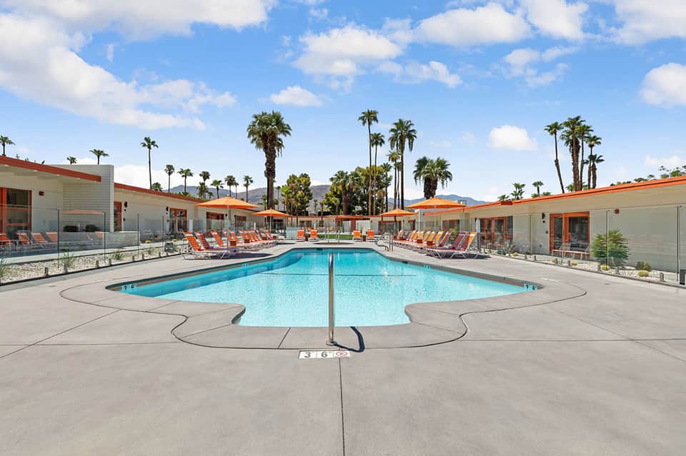 Resort pool with crystal-blue water, orange lounge chairs, palm trees, and mid-century modern buildings