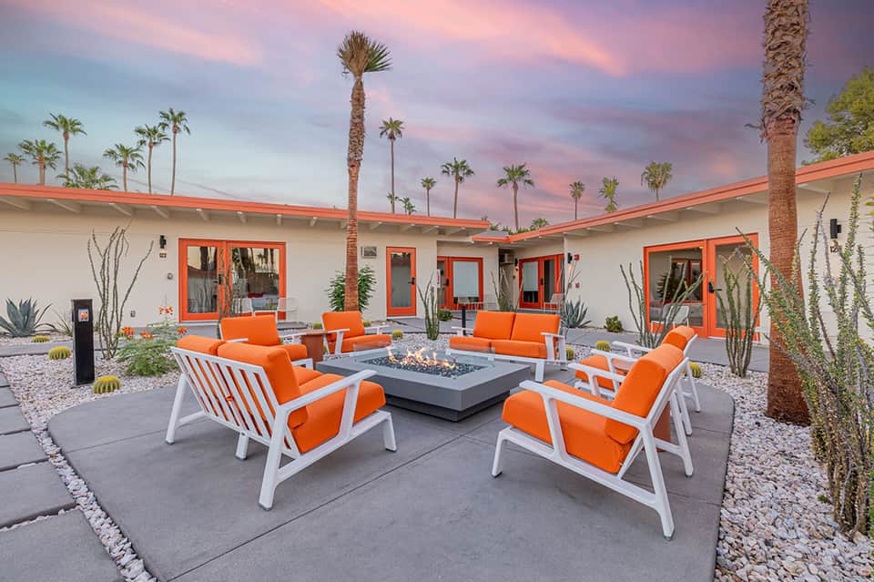 Modern desert courtyard at sunset with orange loungers, fire pit, palm trees, and terracotta roof