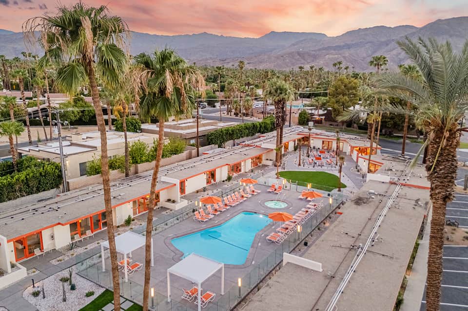 Aerial view of resort pool with orange loungers, spa building, palm trees, and mountain backdrop at sunset