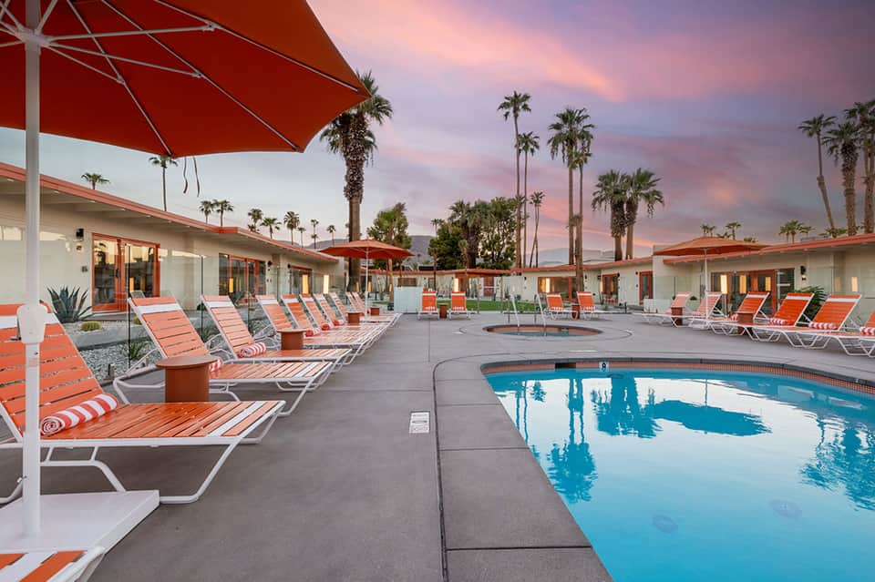 Resort pool at sunset with orange umbrellas, palm trees, and pink-tinged sky above lounge chairs