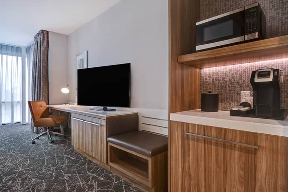 Hotel suite with kitchenette, wooden cabinetry, TV on stand, work desk, and brown leather seating