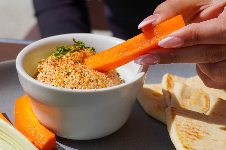 Hand dipping orange carrot strip into hummus dip with crackers nearby