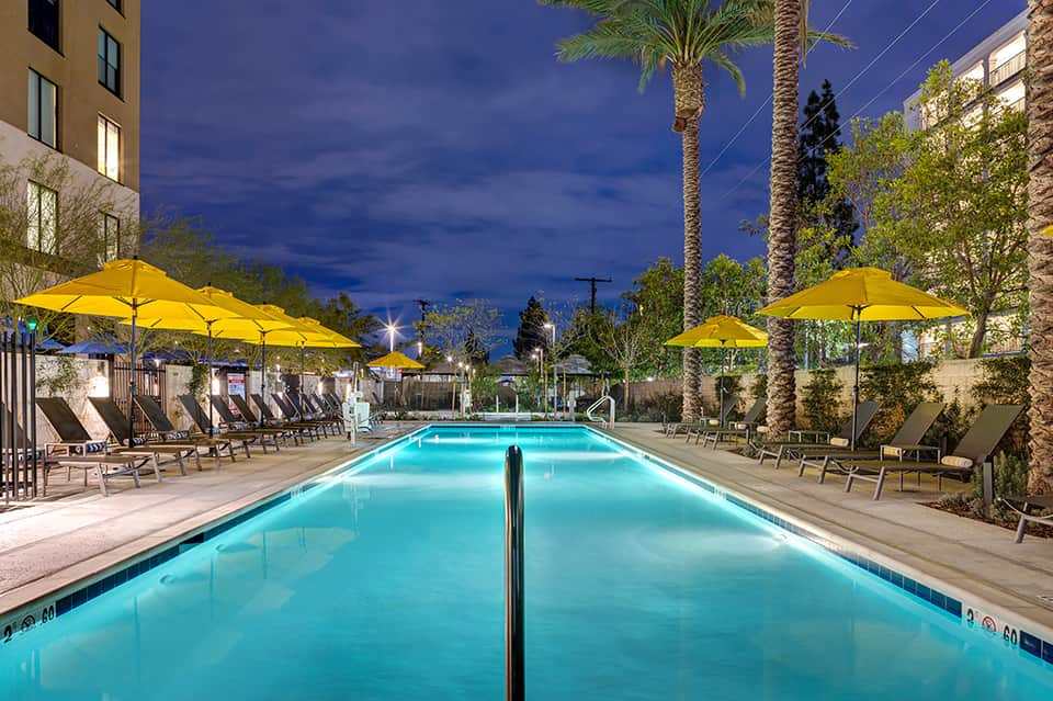 Lit swimming pool at dusk with yellow umbrellas, lounge chairs, and palm trees in courtyard setting