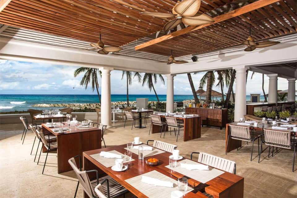 Beachfront dining pavilion with ocean view, wooden ceiling fans, and set tables under pergola