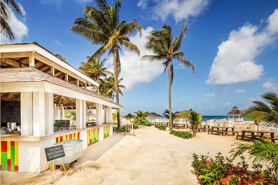 Beachfront bar and restaurant with white structure, palm trees, sand, and ocean view with gazebo