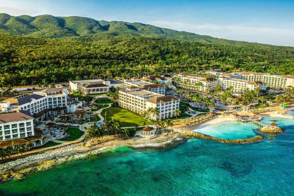 Aerial view of beachfront resort with multiple buildings, lush mountains, turquoise water, and palm trees