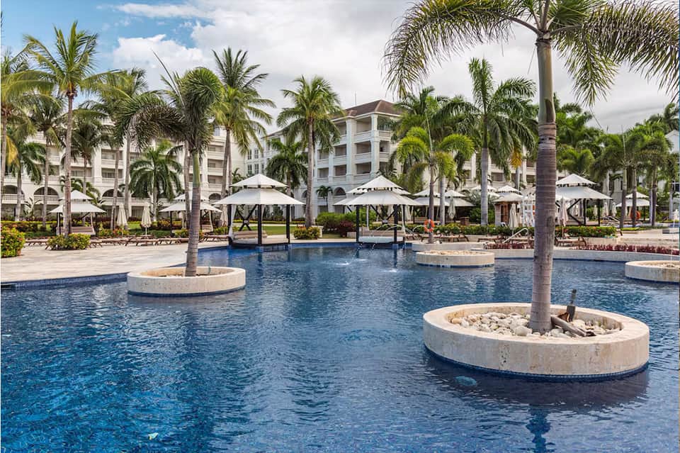 Resort pool with palm trees, white umbrellas, multi-story white hotel building, and lush landscaping