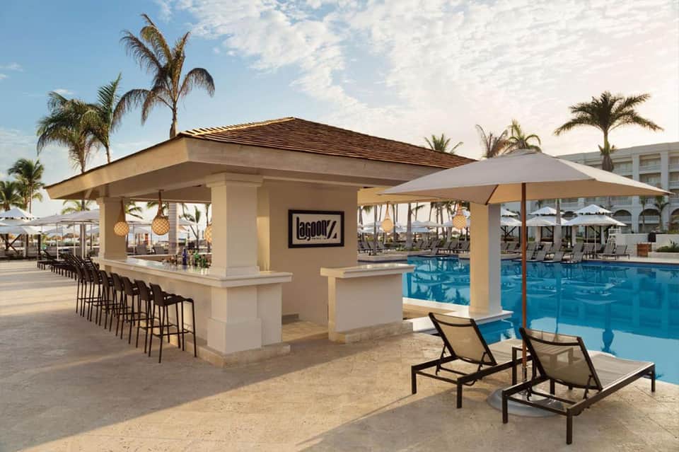 Beachfront pool bar with lounge chairs, white columns, and palm trees under sunny sky
