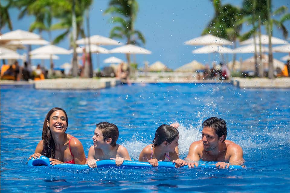 Group of four people enjoying resort pool with white umbrellas and palm trees in background under blue sky
