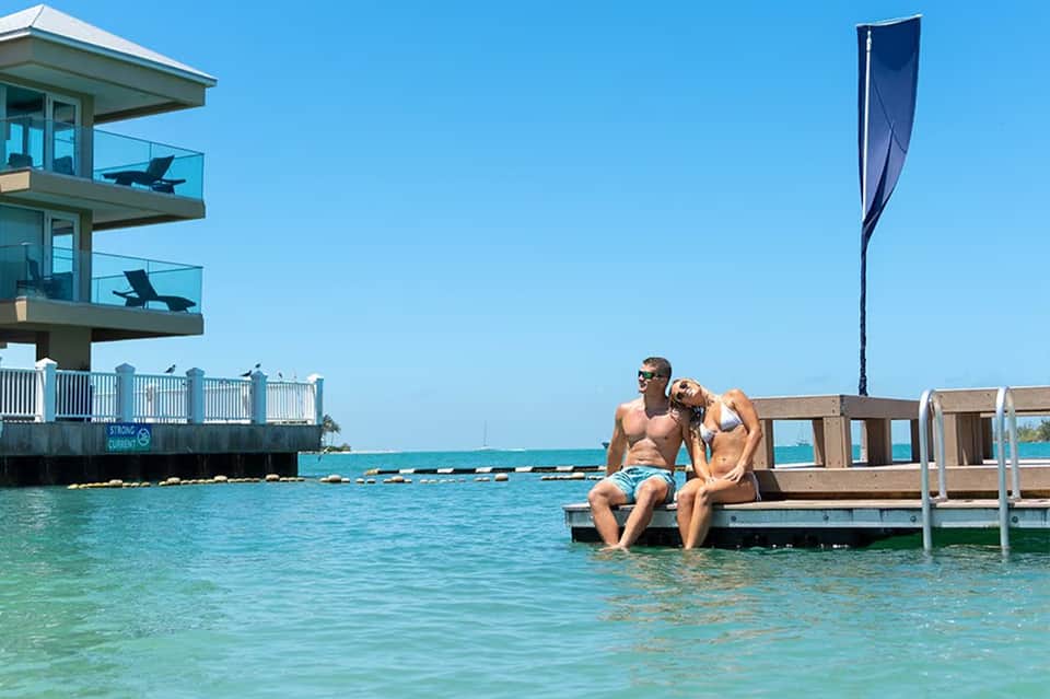 Couple sitting on wooden dock over clear turquoise water with modern waterfront building