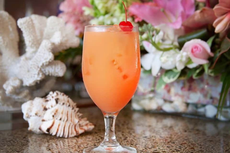 Coral-colored cocktail with cherry garnish surrounded by white seashells and pink flowers